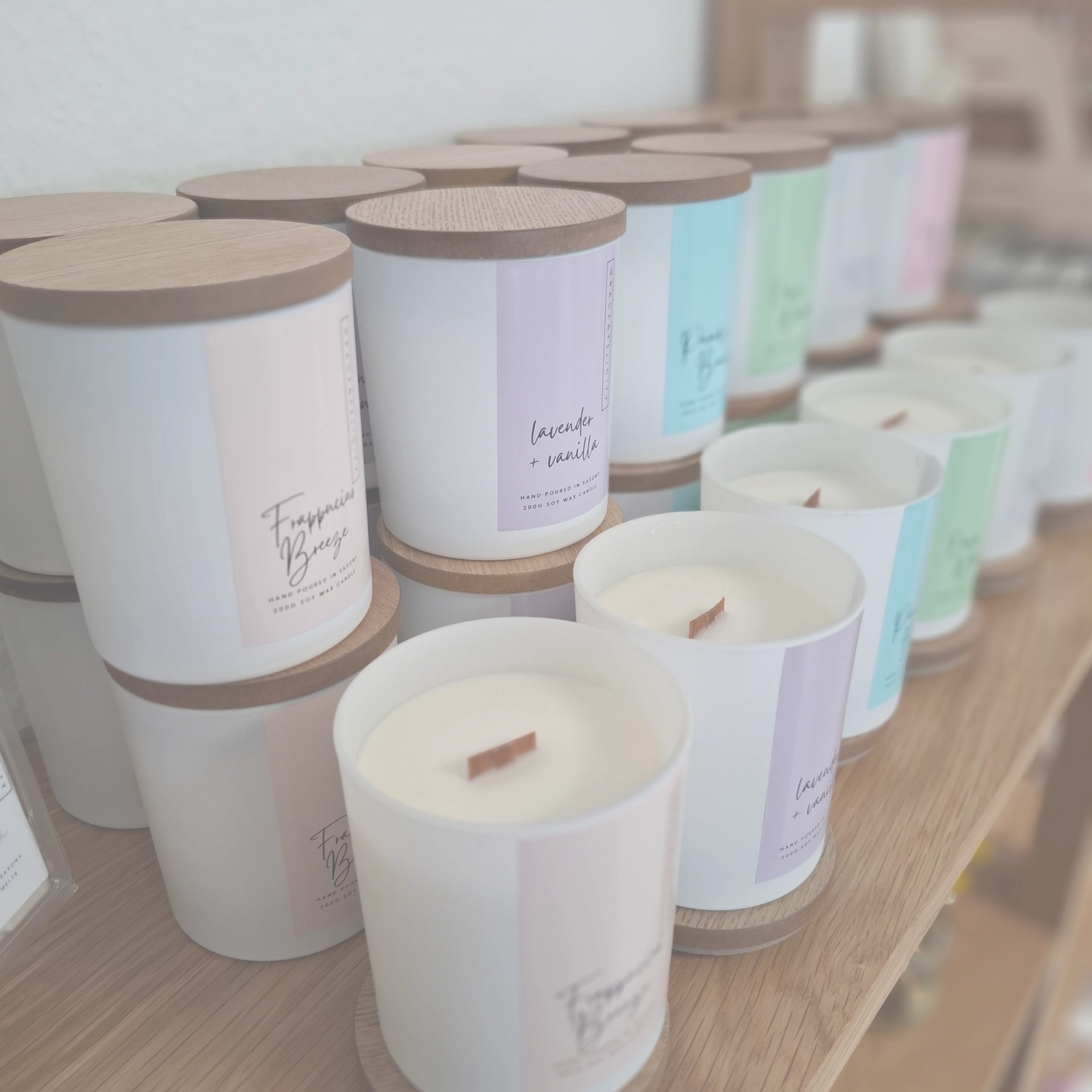 Grace&Felicity - concept store - Eine Reihe großer, weißer Keramikkerzen mit hellen Holzdeckeln und Etiketten in verschiedenen Pastellfarben wie Blau, Lila und Grün.